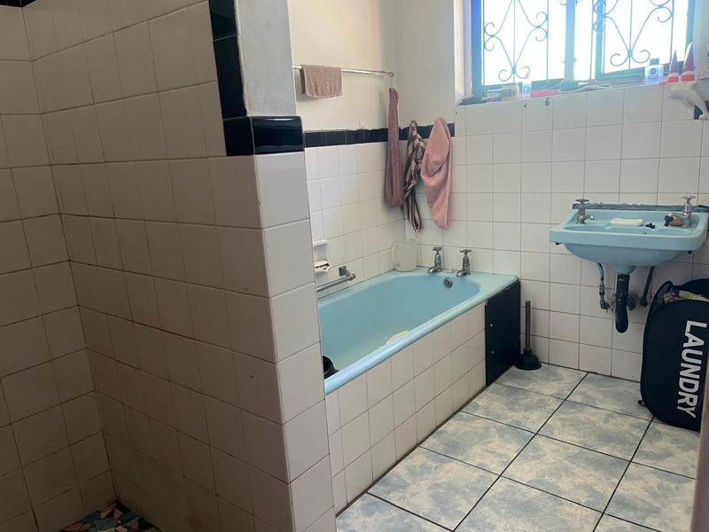A dated bathroom featuring a blue bathtub and matching basin, tiled walls and flooring, and a window with security bars.