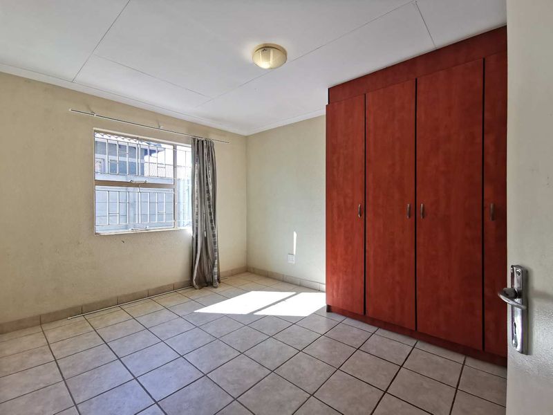A functional, empty bedroom featuring tiled flooring, built-in wooden cupboards, and a window fitted with security bars.