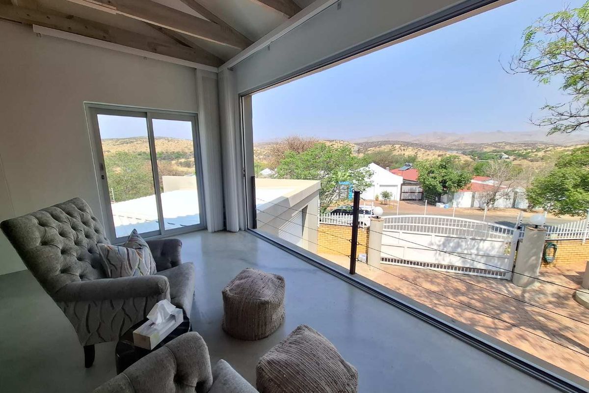 A bright living area featuring polished concrete floors, exposed timber ceiling beams, and expansive floor-to-ceiling glass windows offering panoramic landscape views.