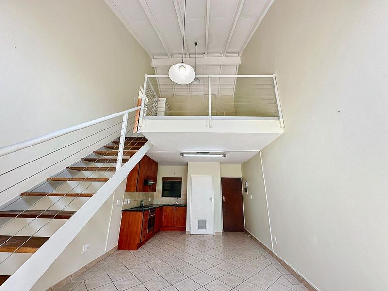 Open-plan living area featuring a mezzanine level, staircase, and a functional kitchen with built-in cabinetry and appliances, set under high exposed beam ceilings.