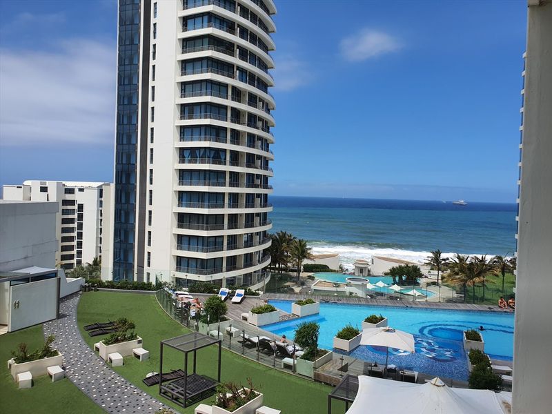 Elevated view overlooking a modern apartment complex, swimming pool area, landscaped gardens, and the ocean coastline.