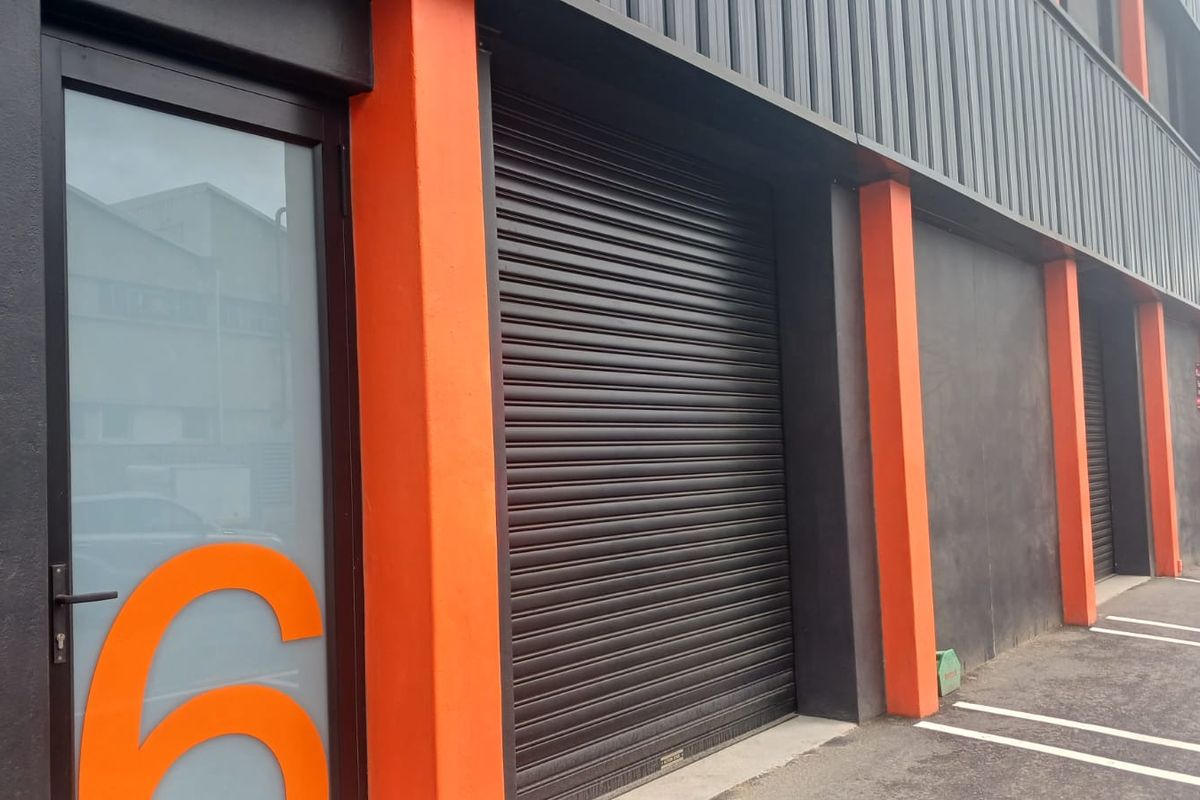 Modern industrial unit exterior featuring a secure roller shutter door, glass entrance, and dedicated parking bay with contemporary orange and dark grey facade.