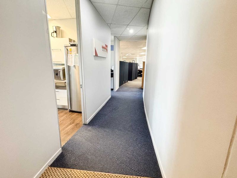 A functional office hallway featuring patterned carpet flooring and a suspended ceiling, providing access to adjacent workspace and kitchen areas.