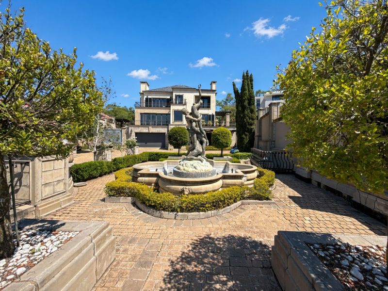 A beautifully landscaped garden featuring a central stone fountain, paved pathways, and manicured hedges, set against a multi-story luxury residence.