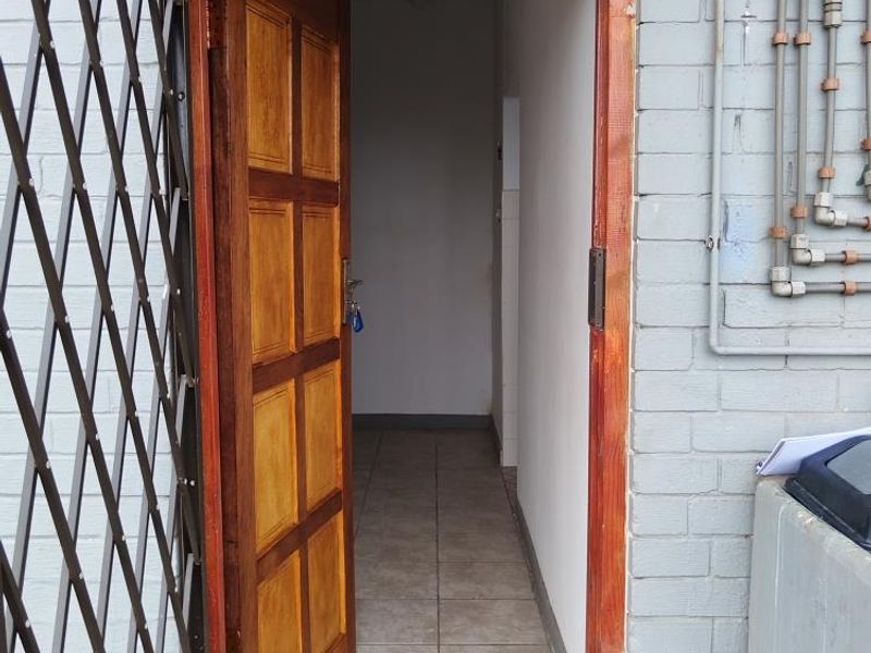 Exterior view of a cottage entrance featuring a timber door, security gate, and tiled flooring with exposed exterior plumbing on the brick facade.
