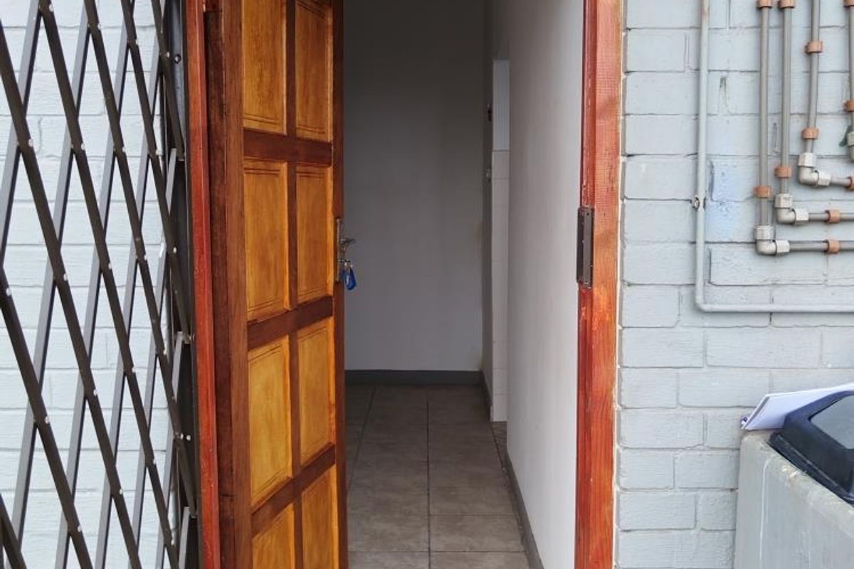 Exterior view of a cottage entrance featuring a timber door, security gate, and tiled flooring with exposed exterior plumbing on the brick facade.