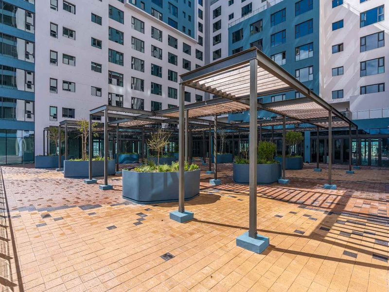 A spacious, modern outdoor terrace featuring paved flooring, metal pergola structures, and integrated planters, set against a contemporary apartment building facade.