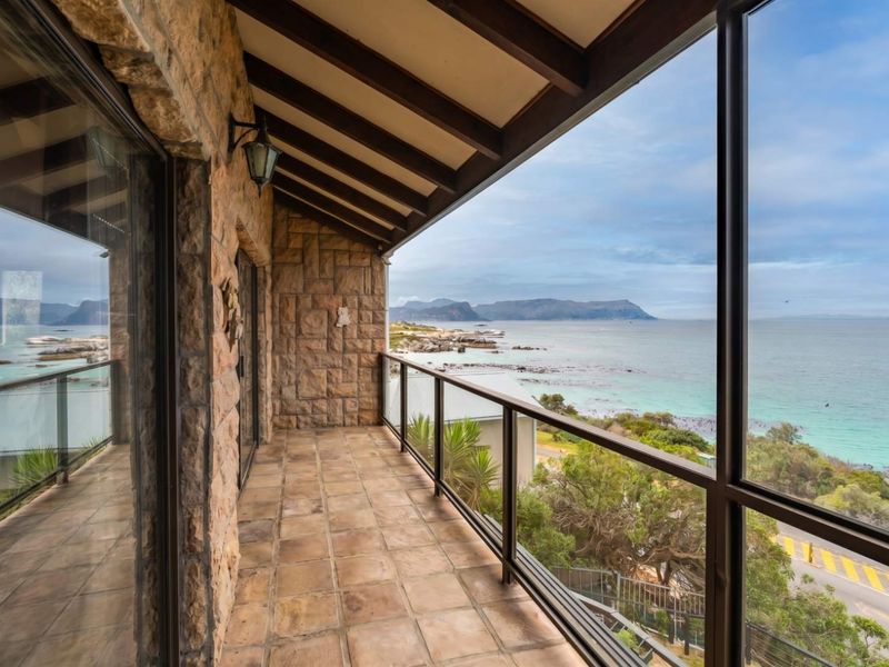 A spacious covered balcony featuring stone-clad walls, exposed timber beams, and a glass balustrade, offering panoramic ocean and coastal views.