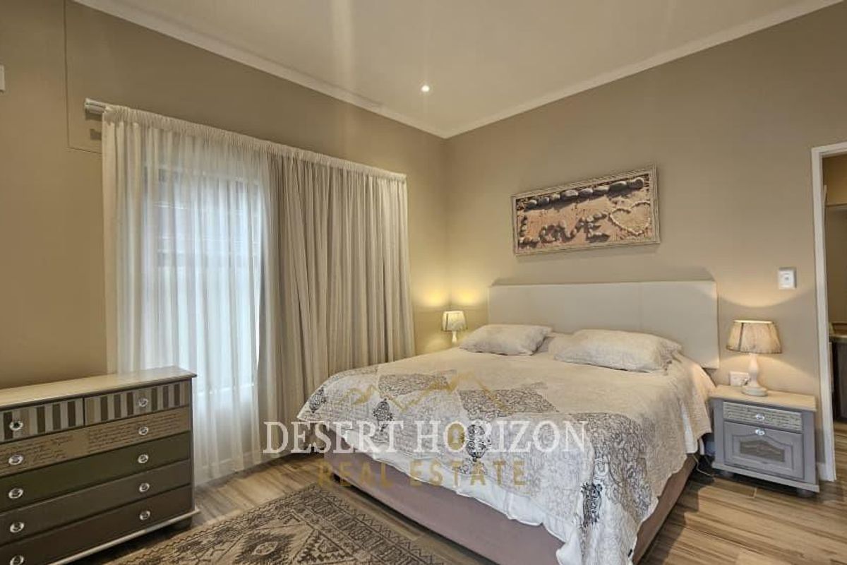 A well-maintained bedroom featuring neutral wall tones, wood-look flooring, and a large window with sheer curtains, providing a bright and comfortable living space.