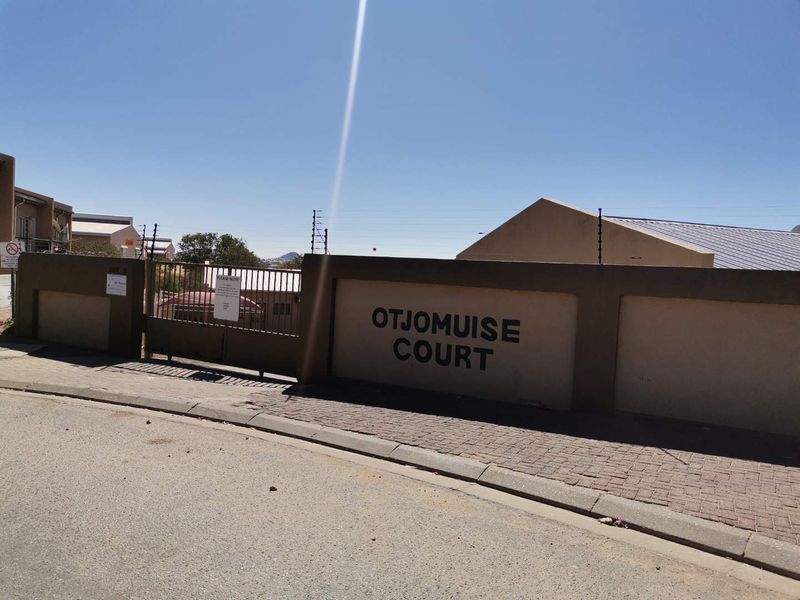 Street view of the Otjomuise Court townhouse complex entrance, featuring a perimeter wall, electric fencing, and a secure metal gate.