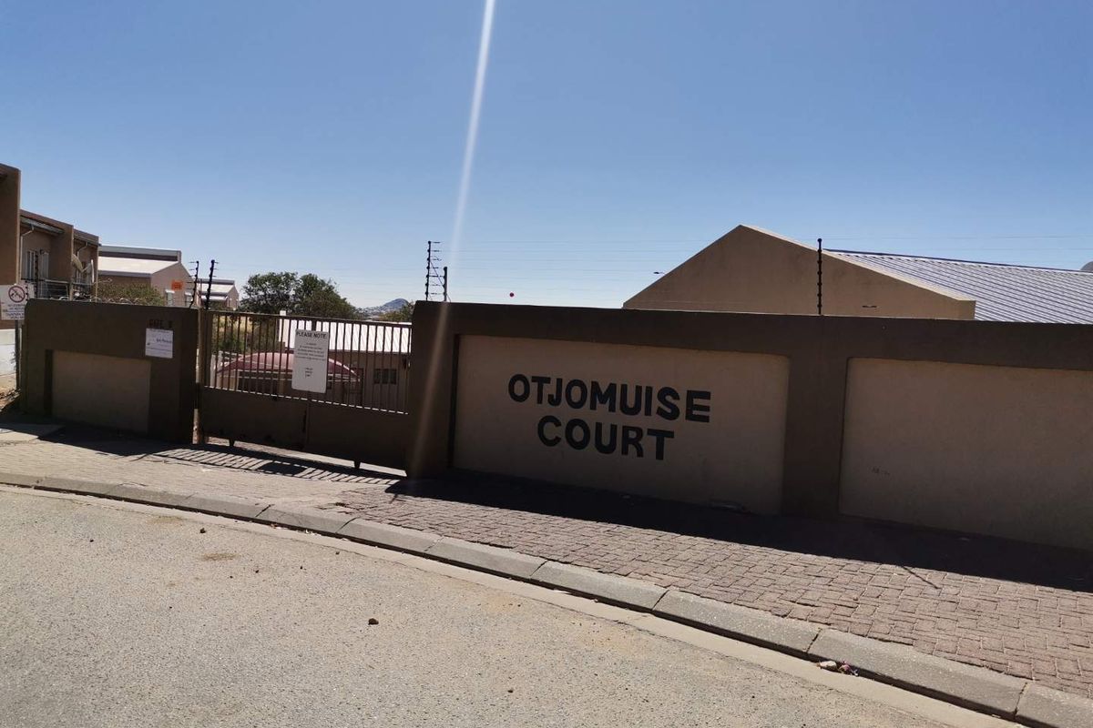 Street view of the Otjomuise Court townhouse complex entrance, featuring a perimeter wall, electric fencing, and a secure metal gate.