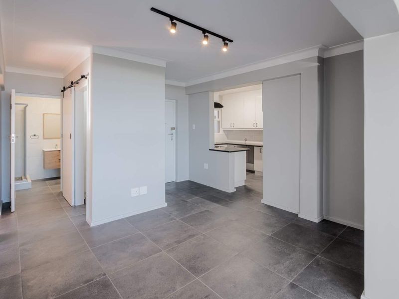 Modern open-plan living area featuring large format floor tiles, track lighting, and a seamless transition to a contemporary kitchen and adjacent bathroom.