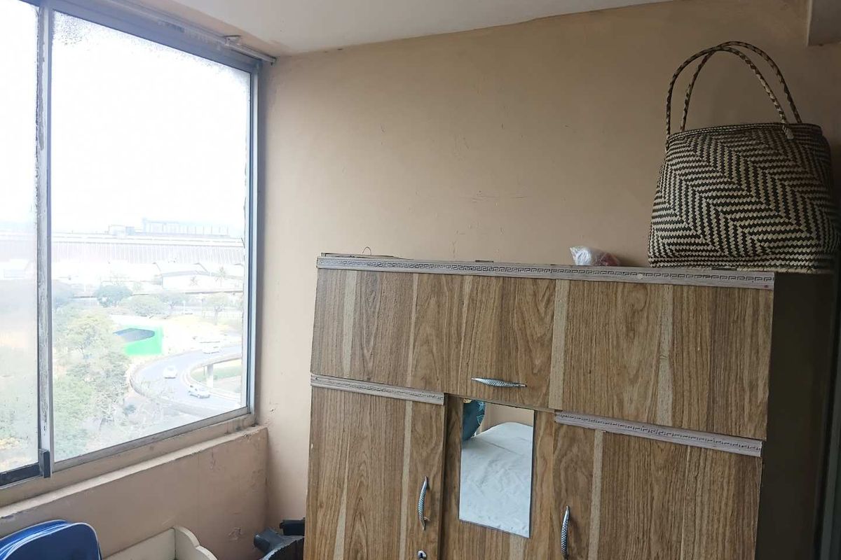 A bedroom featuring a large window with an urban view, showing signs of wear on the walls and window frame.