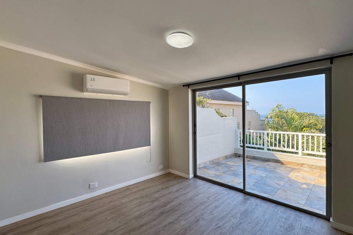 Bright bedroom featuring laminate flooring, a split-system air conditioner, and large sliding glass doors opening onto a private tiled balcony with coastal views.