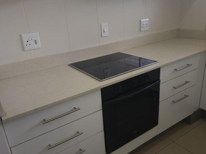 A clean, modern kitchen featuring white cabinetry, stone countertops, a built-in oven, electric hob, and a tiled backsplash.