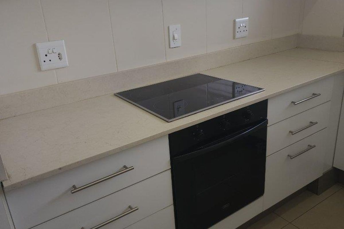 A clean, modern kitchen featuring white cabinetry, stone countertops, a built-in oven, electric hob, and a tiled backsplash.