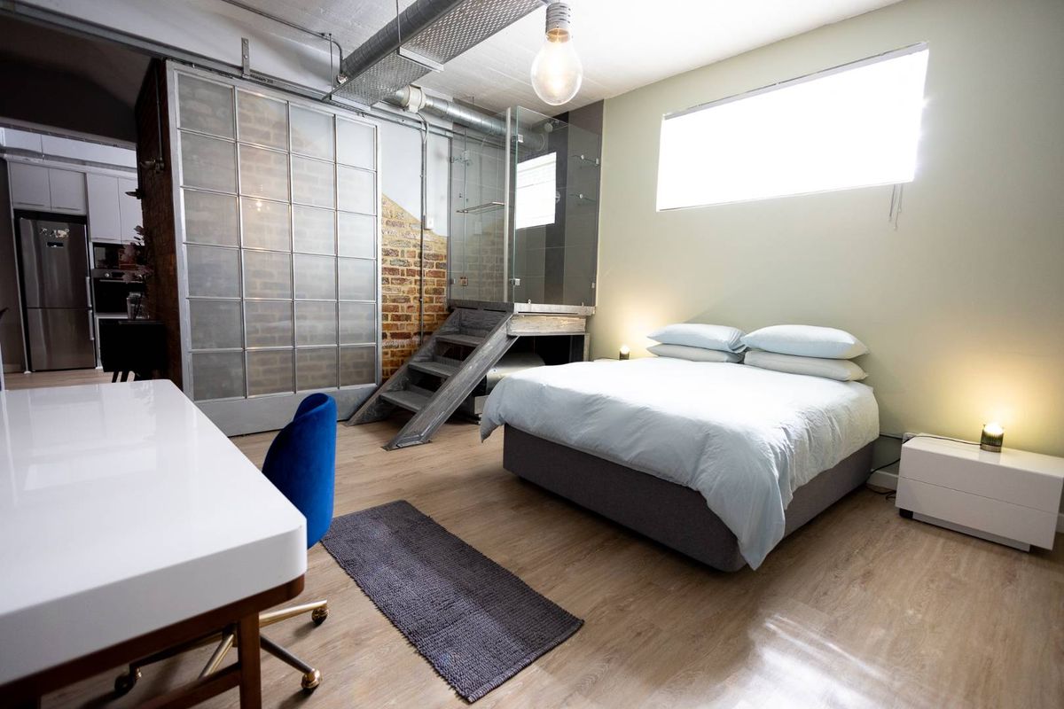 Modern industrial-style bedroom featuring exposed brickwork, high ceilings with exposed ducting, laminate flooring, and an integrated glass-enclosed bathroom area.