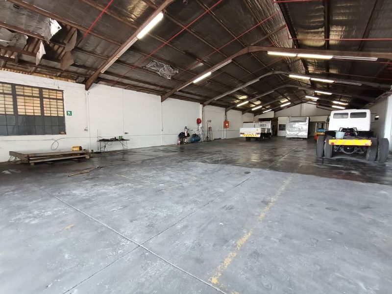 Spacious industrial warehouse featuring high ceilings with exposed steel trusses, concrete flooring, and large metal-framed windows, suitable for storage or workshop operations.