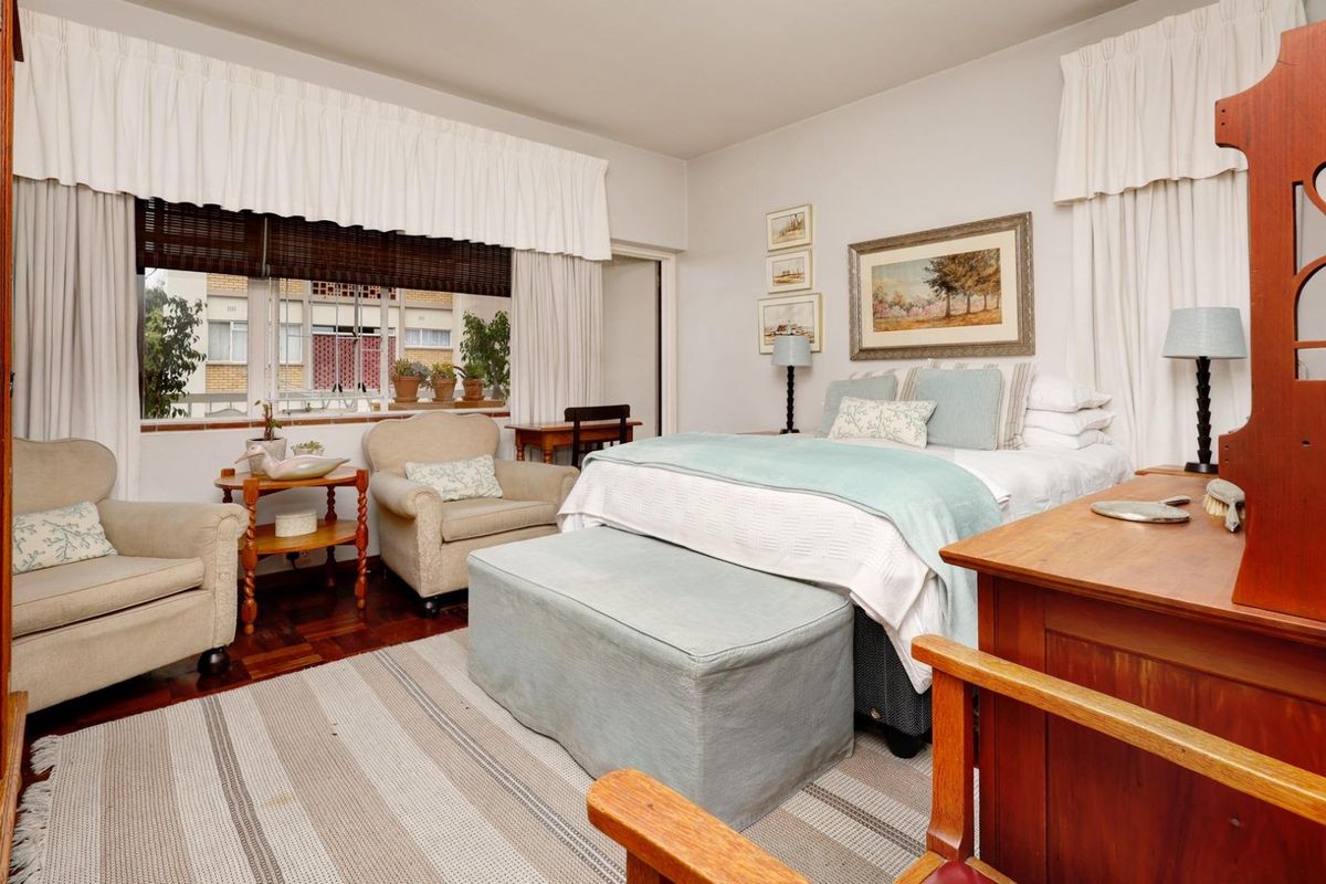 A well-lit bedroom featuring classic parquet flooring and a large window with traditional curtain pelmets, offering a comfortable and functional living space.