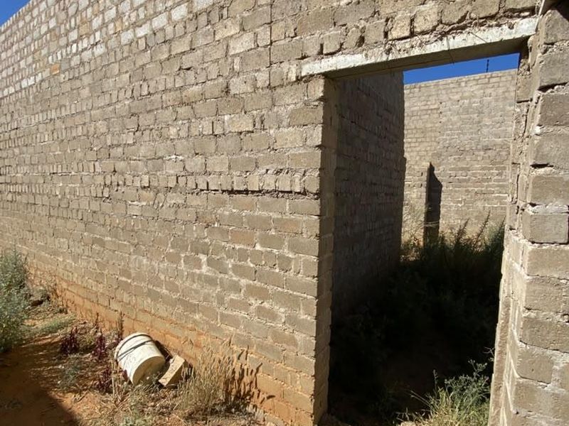 An unfinished residential structure featuring exposed brick masonry walls and a concrete lintel, currently in a shell state with surrounding overgrown vegetation.