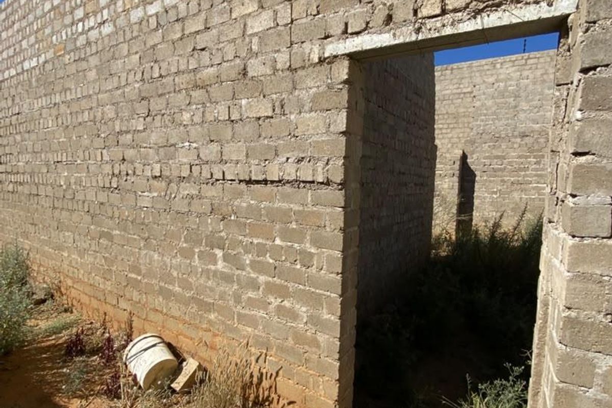 An unfinished residential structure featuring exposed brick masonry walls and a concrete lintel, currently in a shell state with surrounding overgrown vegetation.