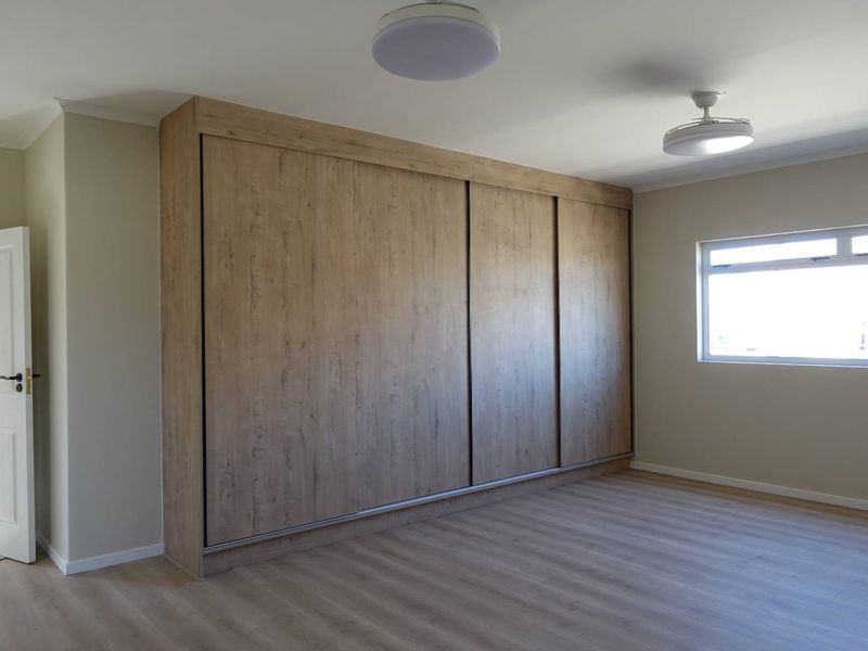 A bright, empty bedroom featuring modern wood-finish built-in sliding door wardrobes, light laminate flooring, and neutral wall tones.