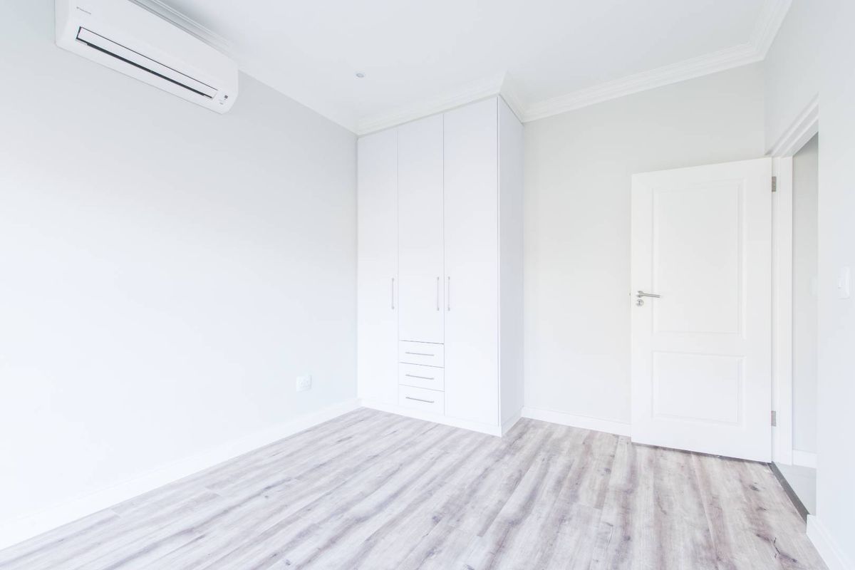 A bright, modern bedroom featuring light wood-look flooring, built-in white cabinetry, and a wall-mounted air conditioning unit.