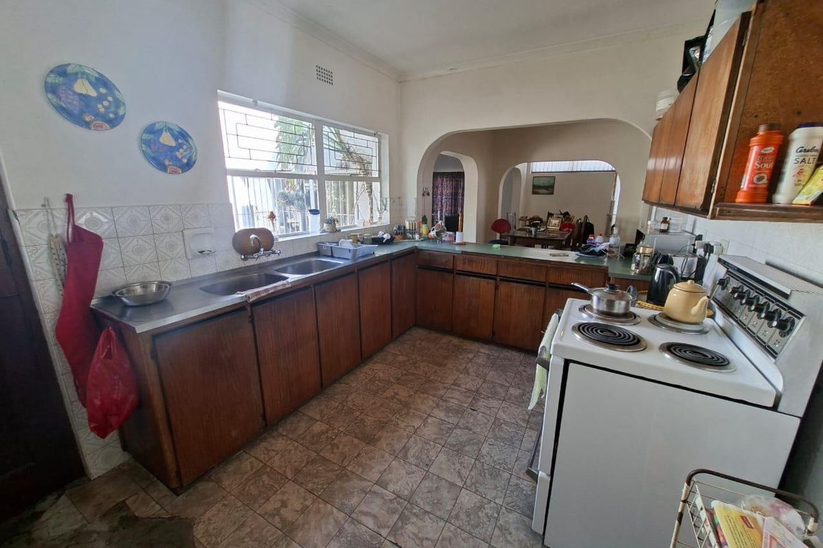 A dated kitchen featuring wooden cabinetry, a double stainless steel sink, and tiled flooring, with an open-plan layout leading into the dining area.