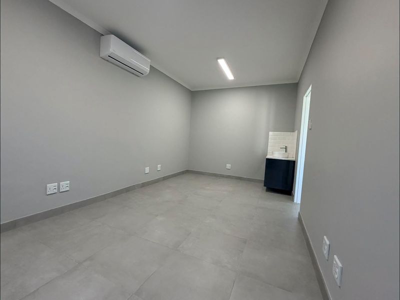 A clean, modern office space featuring neutral grey walls, large format floor tiles, an integrated air conditioning unit, and a convenient built-in vanity basin.