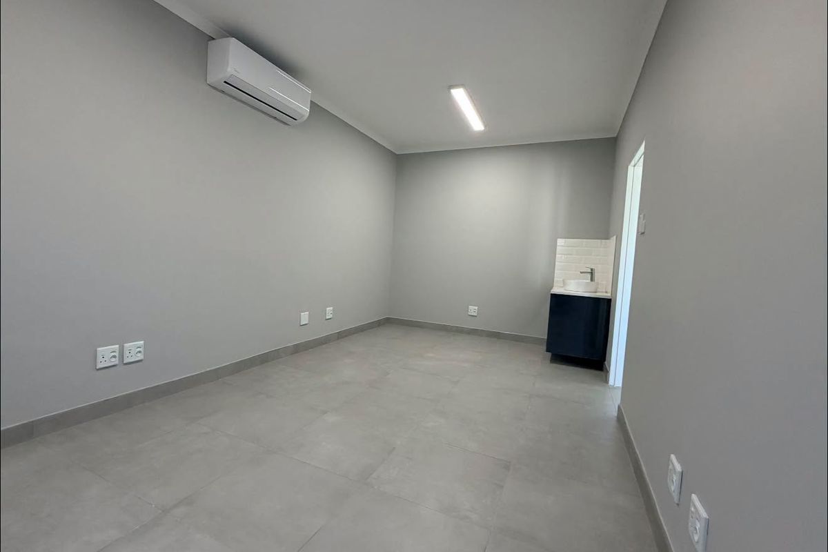 A clean, modern office space featuring neutral grey walls, large format floor tiles, an integrated air conditioning unit, and a convenient built-in vanity basin.