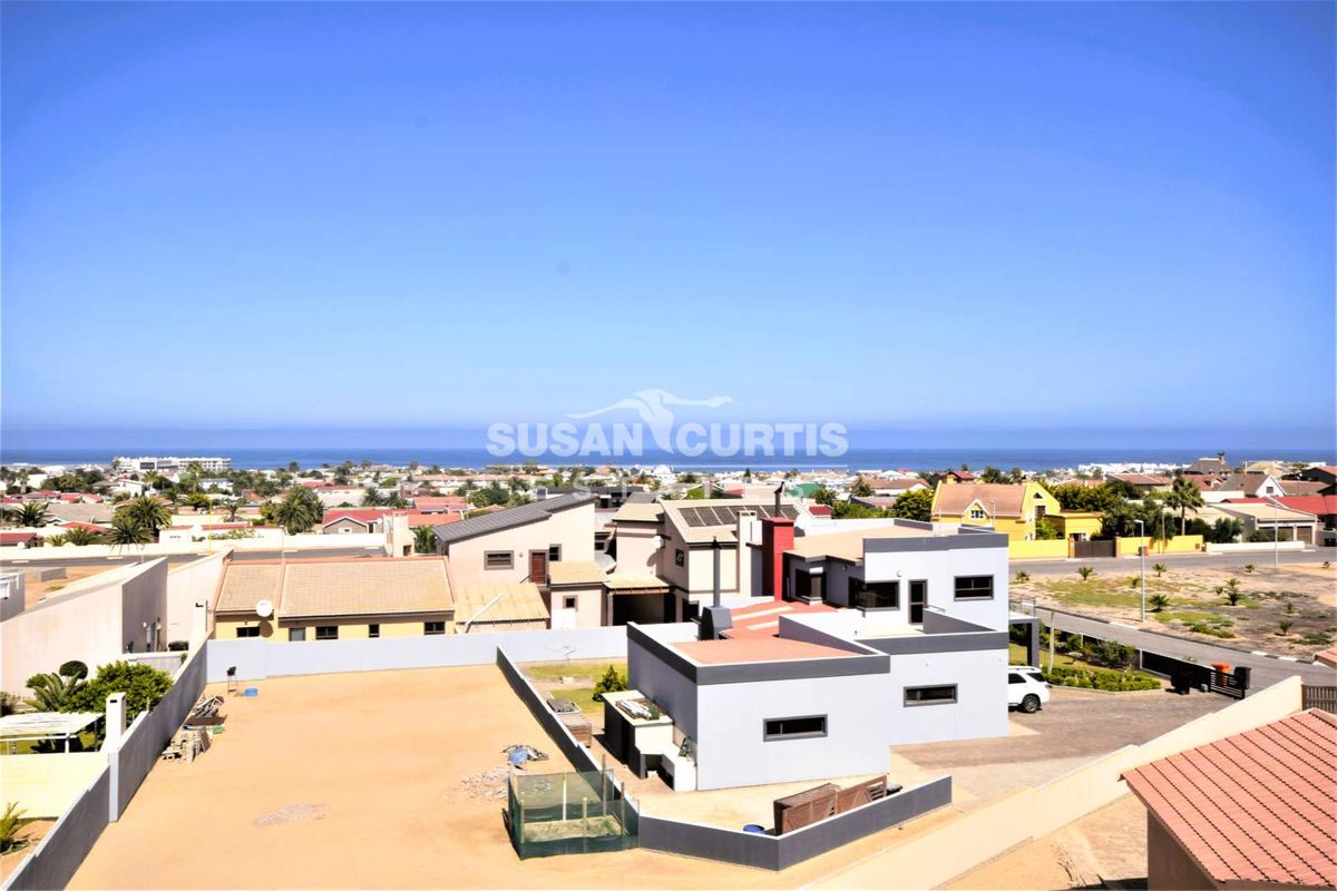 Aerial view of the property showing the modern house structure, solar panels, paved driveway, and surrounding residential neighborhood with ocean views.