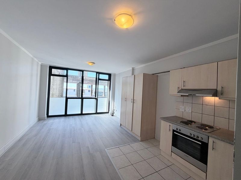 Open-plan living area featuring a compact kitchen with built-in cabinetry, electric oven, and hob, leading to a bright space with large windows.