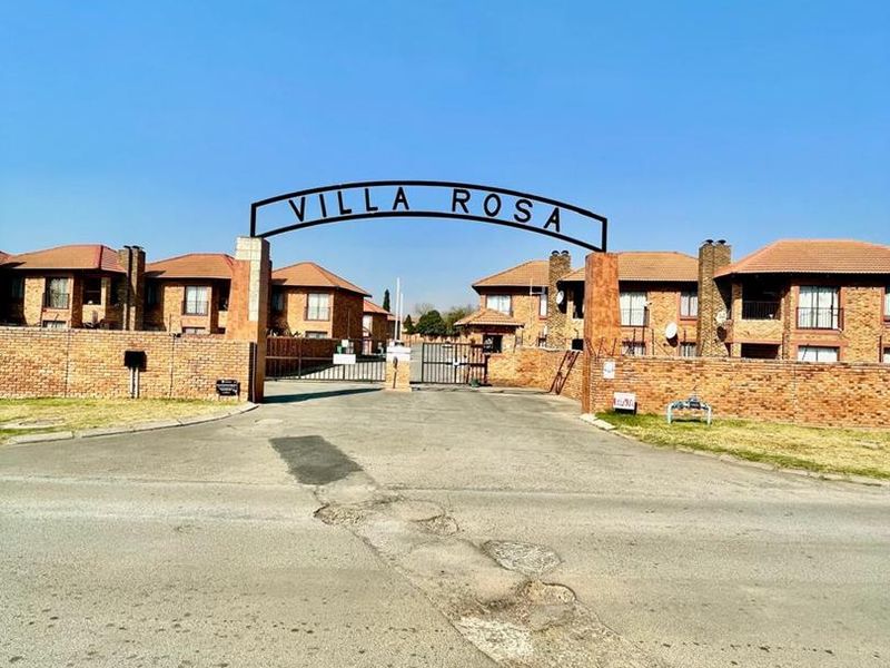 The entrance to Villa Rosa complex featuring a brick boundary wall, metal gate, and paved driveway access.