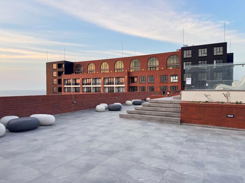 A spacious rooftop terrace featuring modern grey tiling, brick perimeter walls, and a glass balustrade, offering an expansive outdoor area with architectural views.
