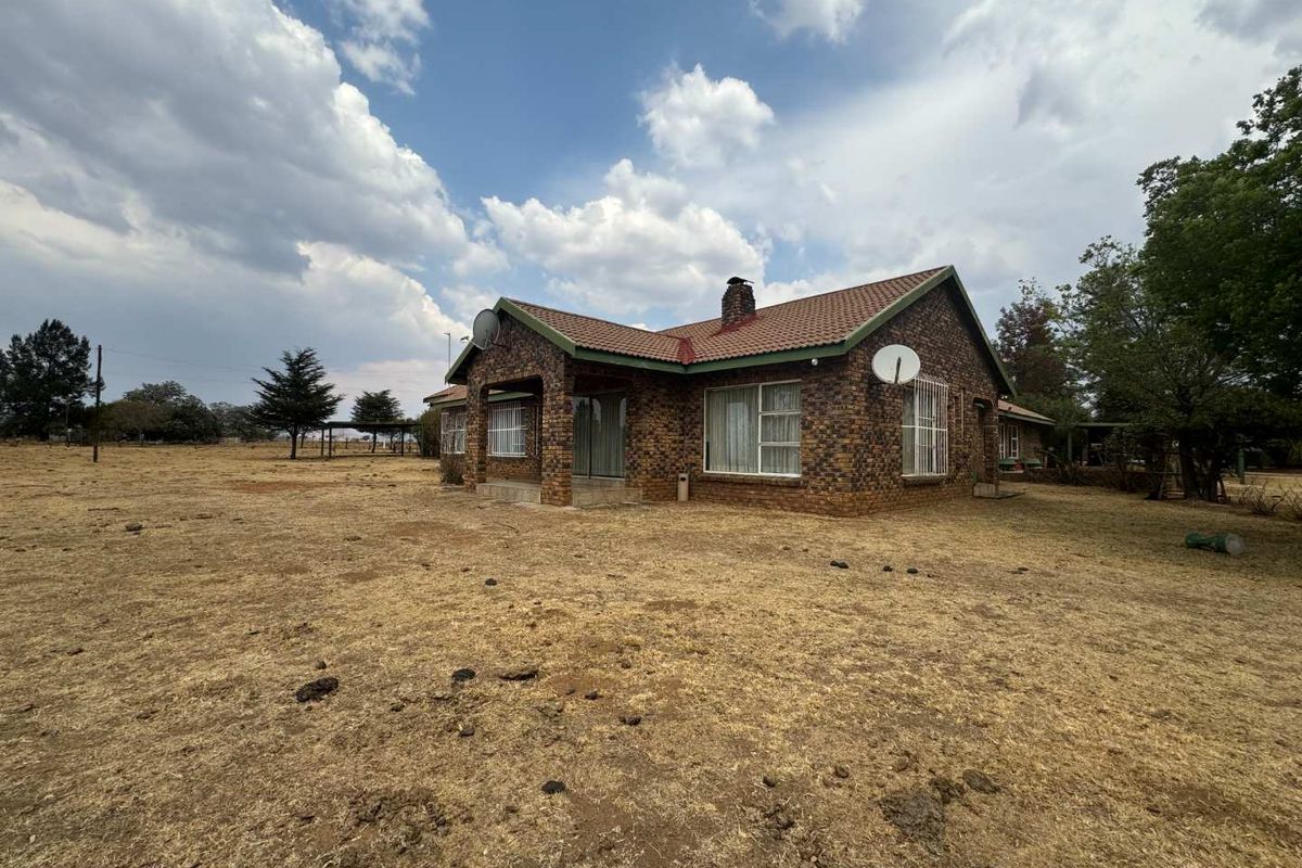 A detached face brick house featuring a tiled roof, chimney, and security bars, set on a large, dry grass plot.