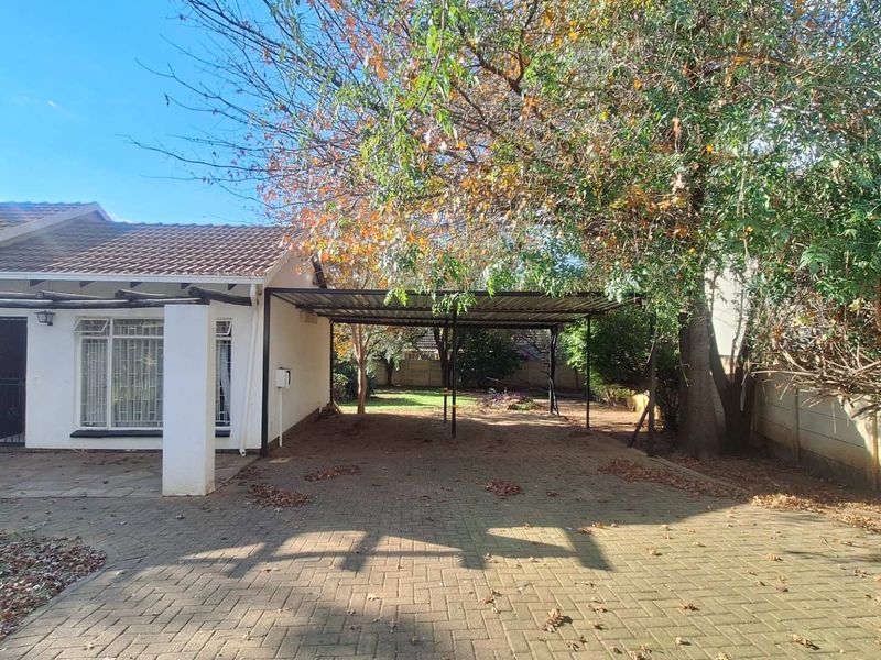 Rear exterior view featuring a paved driveway, a covered carport structure, and a garden area with mature trees and perimeter walling.