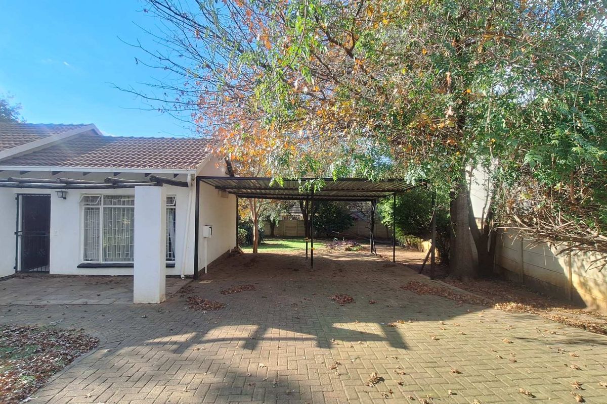 Rear exterior view featuring a paved driveway, a covered carport structure, and a garden area with mature trees and perimeter walling.