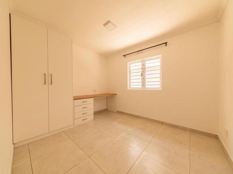 A clean, empty bedroom featuring neutral tiled flooring, a built-in wardrobe, and a functional built-in desk area under a louvered window.