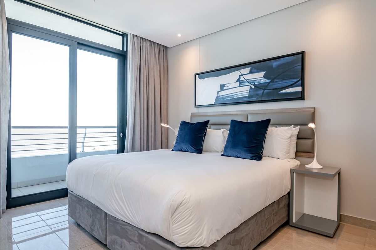 A modern, well-maintained bedroom featuring tiled flooring, recessed ceiling lighting, and large sliding glass doors providing access to a private balcony.