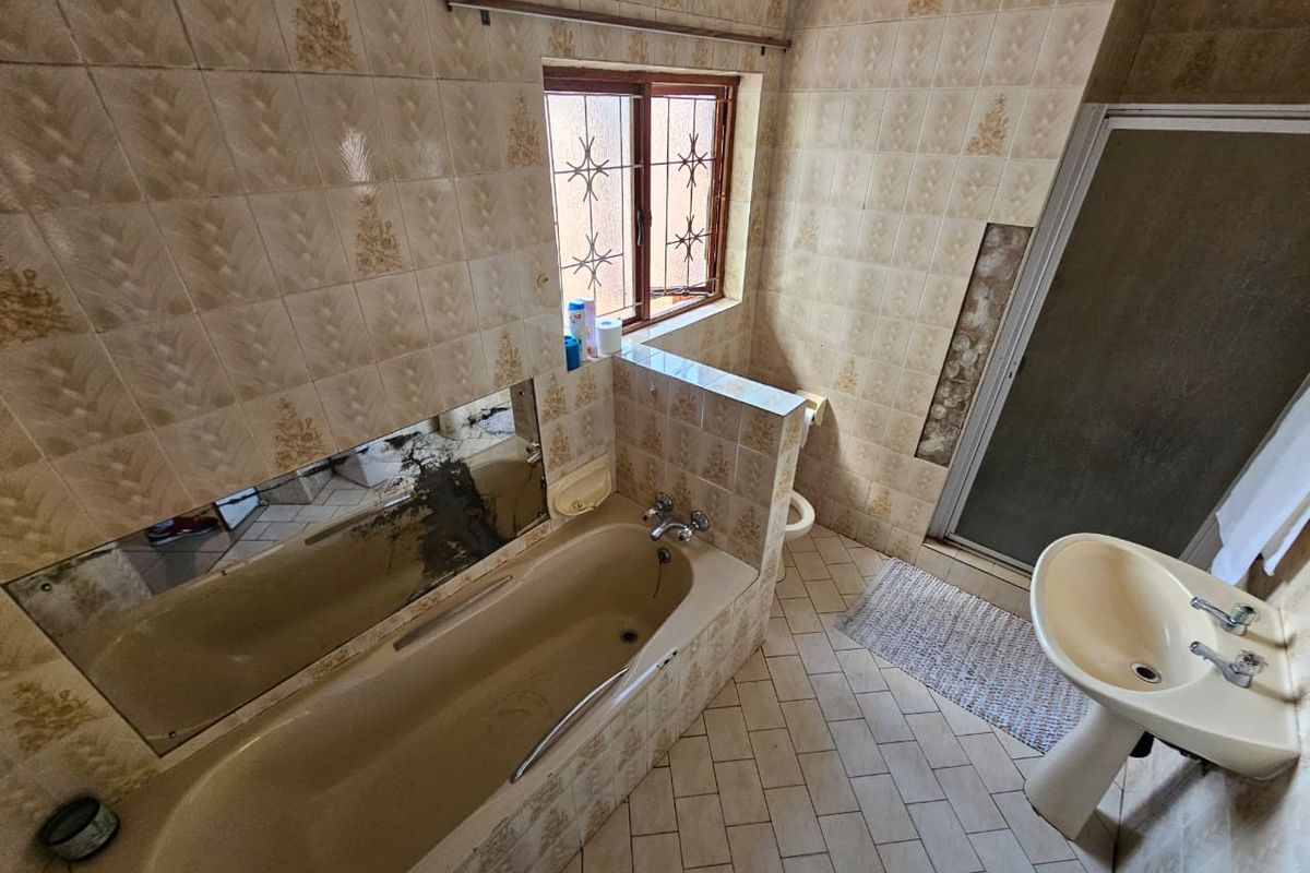 A dated bathroom featuring a built-in bathtub, pedestal sink, and toilet, with significant wear to the wall tiles and mirror.