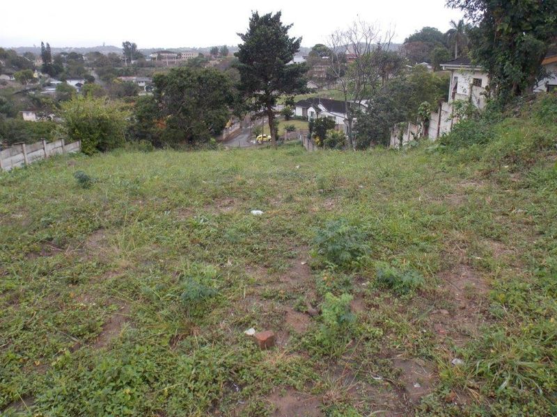 A sloped vacant residential plot featuring established greenery and perimeter retaining walls, offering an elevated view of the surrounding neighbourhood.