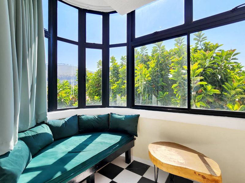 A bright sunroom or enclosed balcony featuring large bay windows with black frames and classic checkered floor tiles, offering a pleasant garden view.