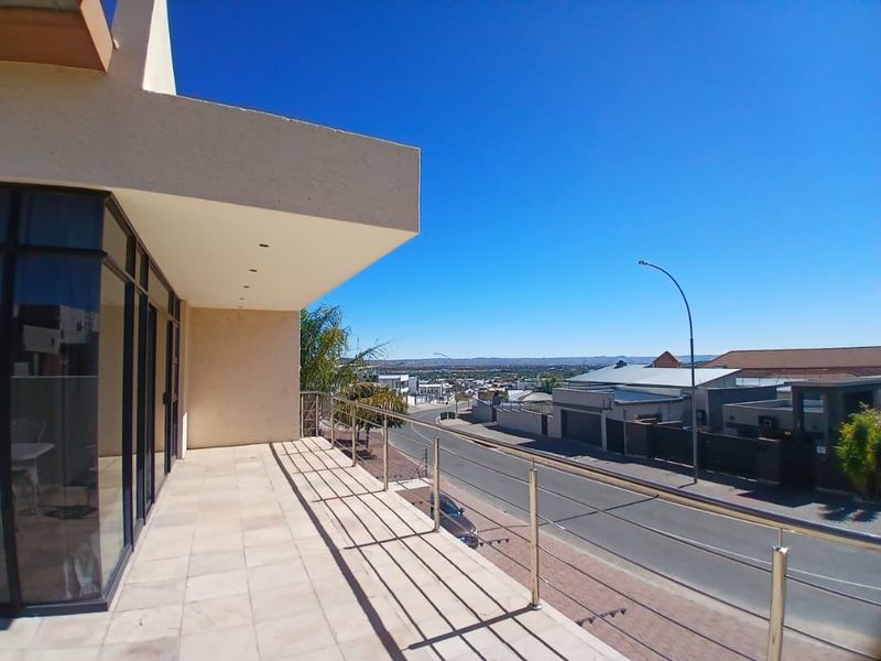 A spacious covered balcony featuring tiled flooring and a modern stainless steel railing, offering an elevated view of the surrounding neighbourhood and street.