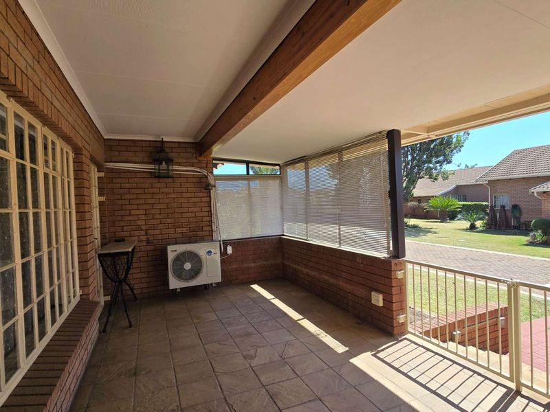 A covered patio area featuring exposed brick walls, tiled flooring, and a metal balustrade, overlooking the surrounding residential garden and driveway.