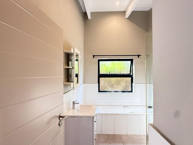 A clean, modern bathroom featuring a built-in vanity, a tiled bathtub, and a glass shower screen under high ceilings with exposed beams.