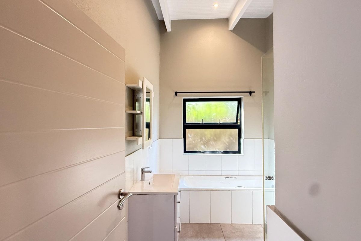 A clean, modern bathroom featuring a built-in vanity, a tiled bathtub, and a glass shower screen under high ceilings with exposed beams.