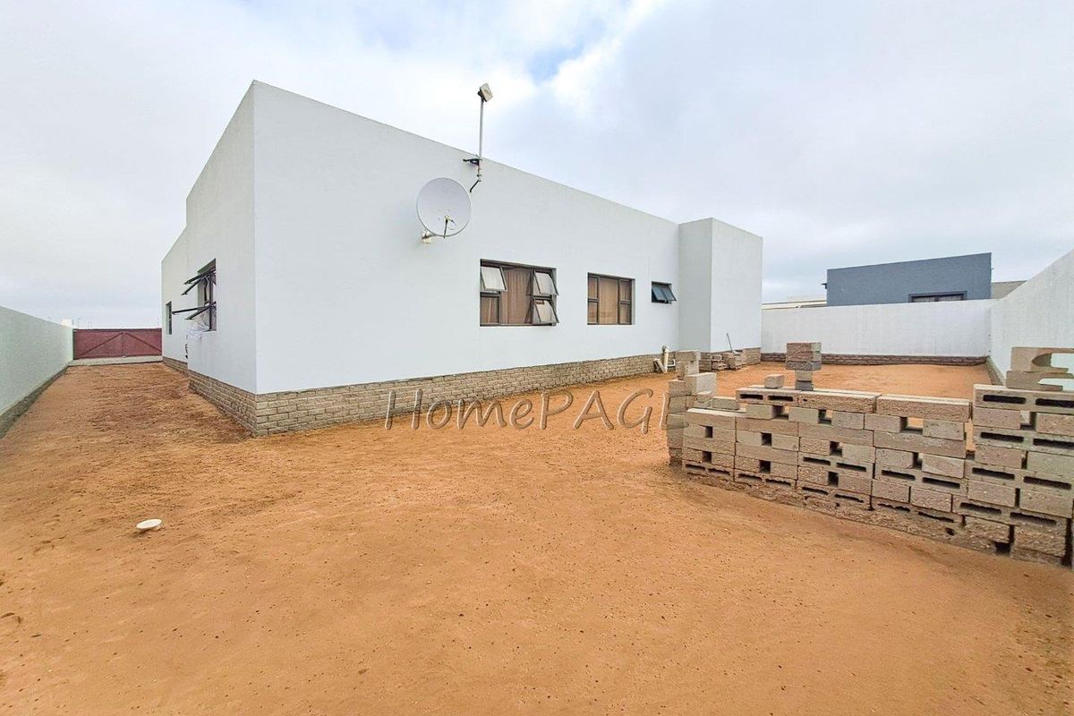 Rear view of the property showing a plain exterior facade, satellite dish, and an unfinished sandy backyard with a low concrete block boundary wall.
