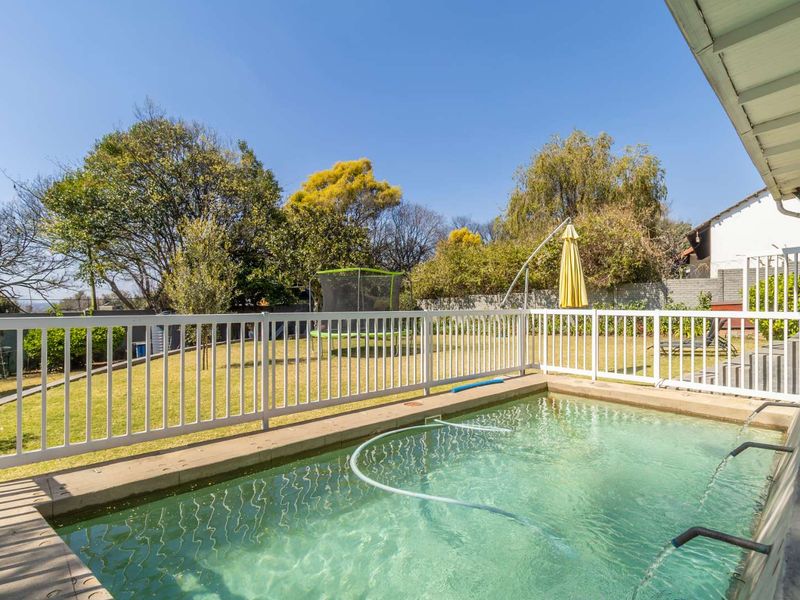 A well-maintained swimming pool area featuring a white safety fence, concrete coping, and a built-in water feature overlooking a spacious garden.
