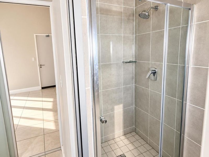 A clean, modern bathroom featuring a glass-enclosed shower with neutral wall tiling and tiled flooring, adjacent to an open hallway area.