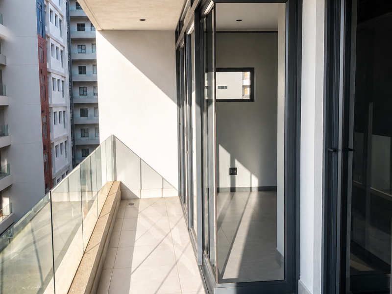 A modern, covered balcony featuring sleek glass balustrades, neutral floor tiles, and large aluminium sliding doors leading into the apartment interior.